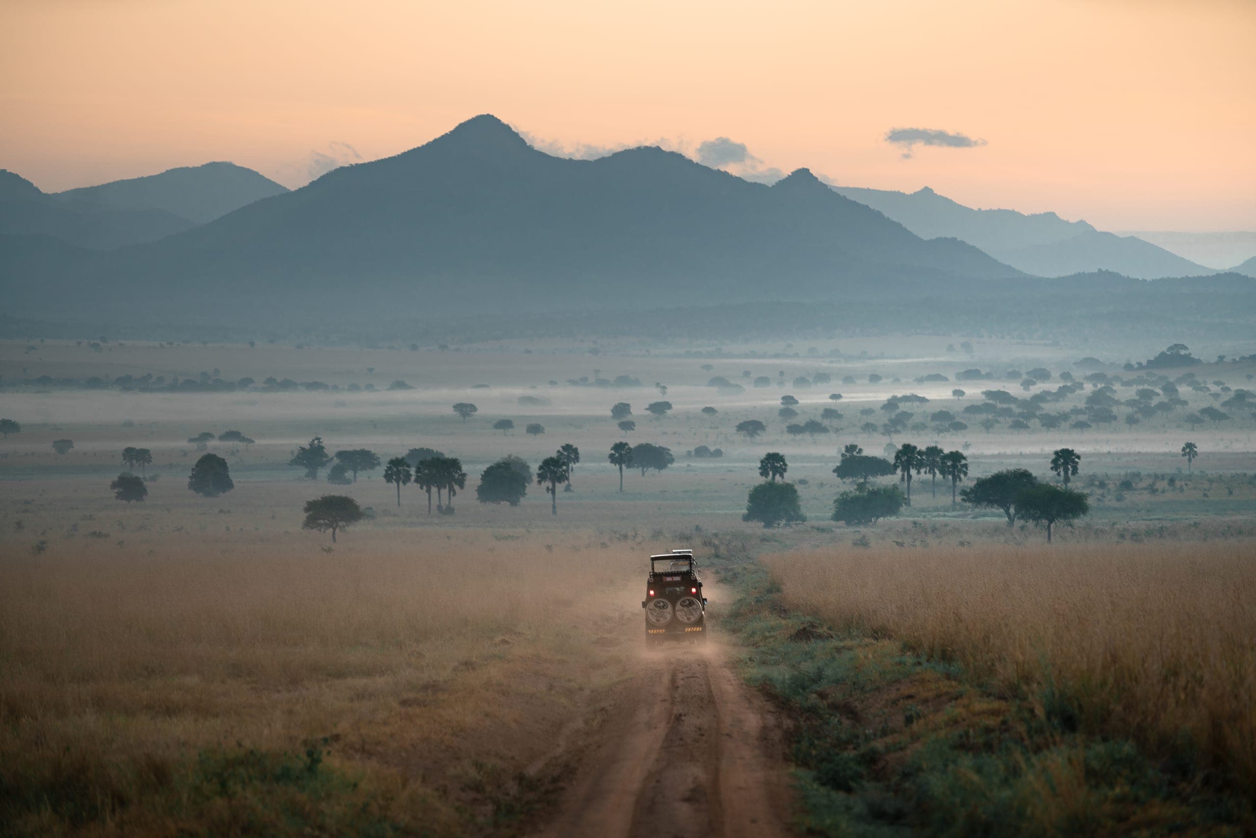 Een safarivoertuig rijdt bij zonsopkomst door Kidepo Valley, met bergen op de achtergrond